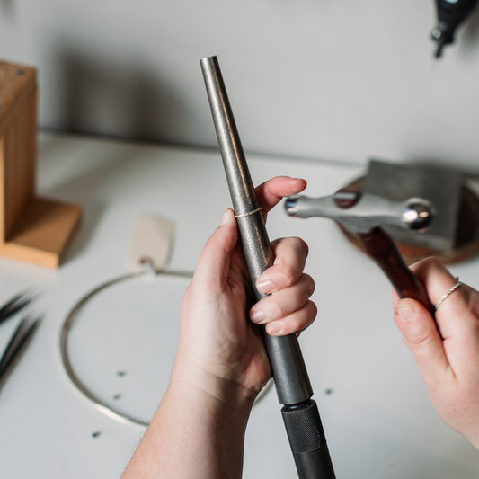 A person's hands holding a metal ring mandrel and hammer with tools in the background on a workshop table.