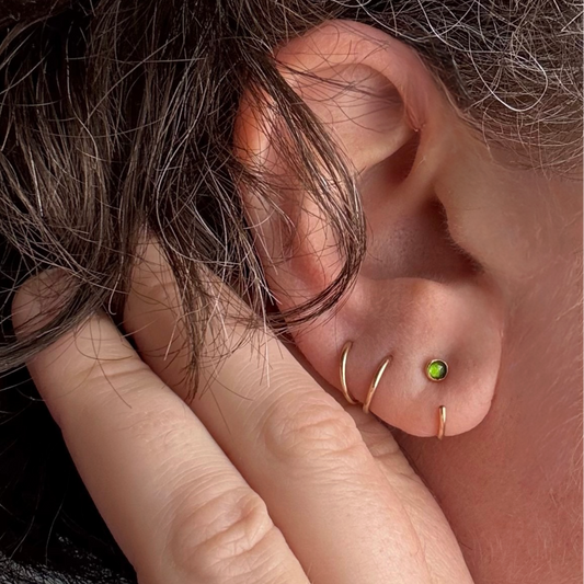 Close-up of a person's ear with multiple gold piercings, including a small green gemstone stud, shown against dark hair and skin.