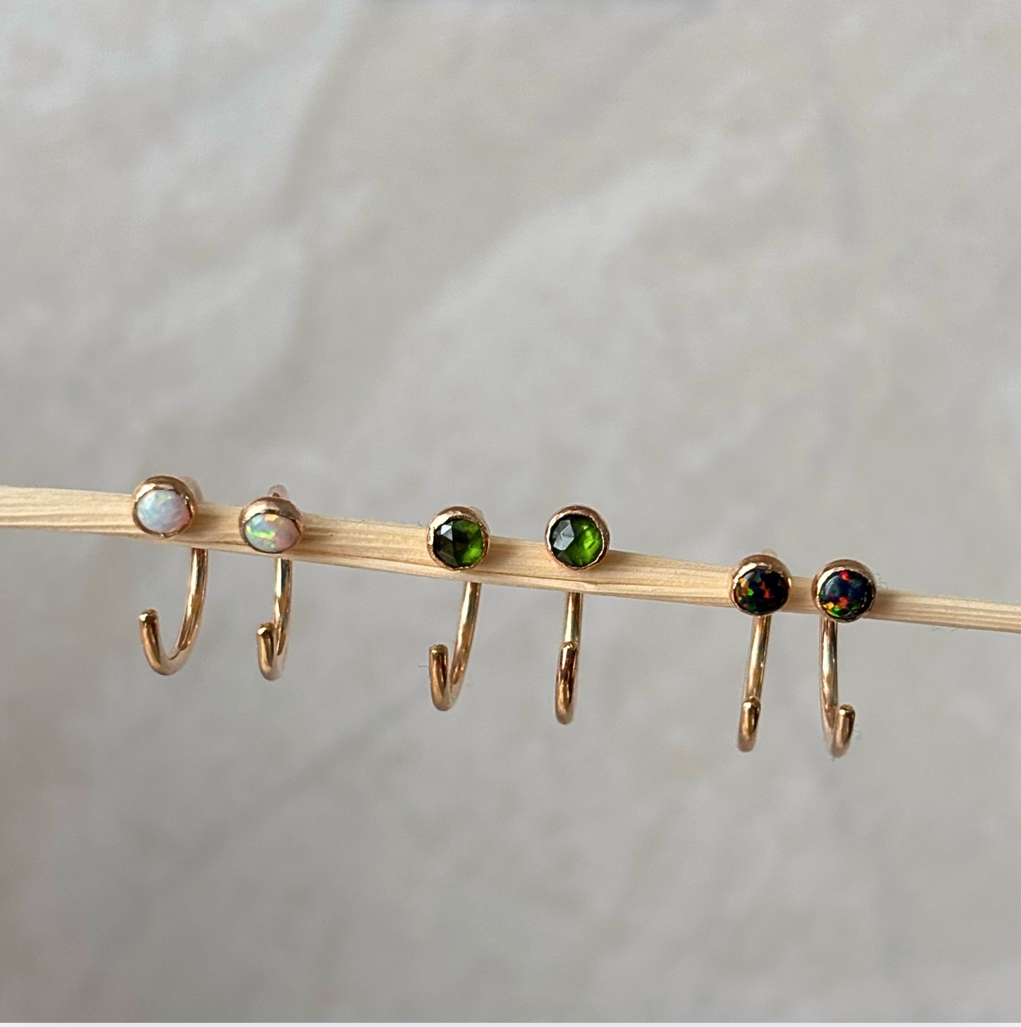 Six gold hoop earrings with round gemstone tops are displayed on a thin wooden stick against a soft light gray background. The stones include white, iridescent opal-like, green, and dark iridescent colors.