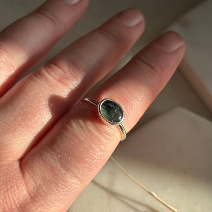Silver ring with a green stone on a person's finger against a neutral background