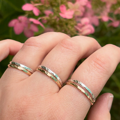 Three slim hammered metal rings in gold, silver, and rose gold tones worn on a person's hand, with a soft-focus background of pink flowers and green foliage.