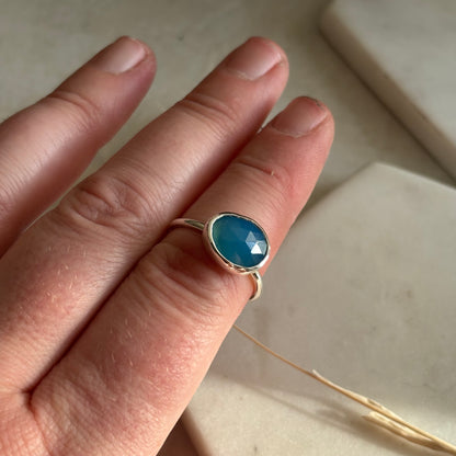 Hand wearing a gold ring with a blue gemstone on a neutral background