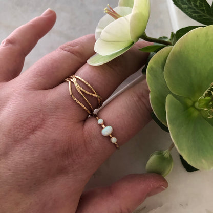 A hand wearing an oval triple gemstone ring with white opal set in gold with flowers in the background.