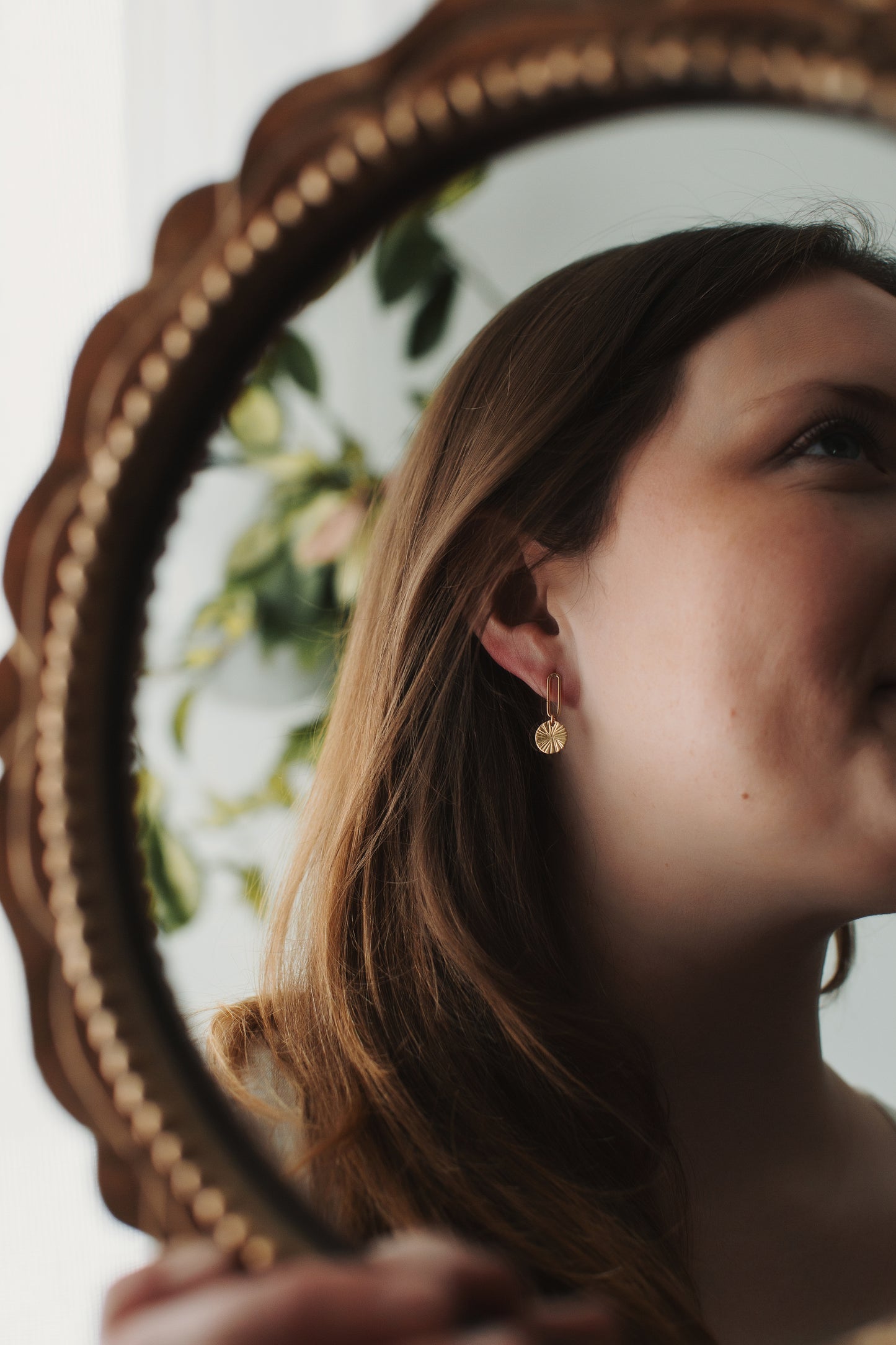 Close-up of a person wearing gold earrings in front of a decorative mirror.