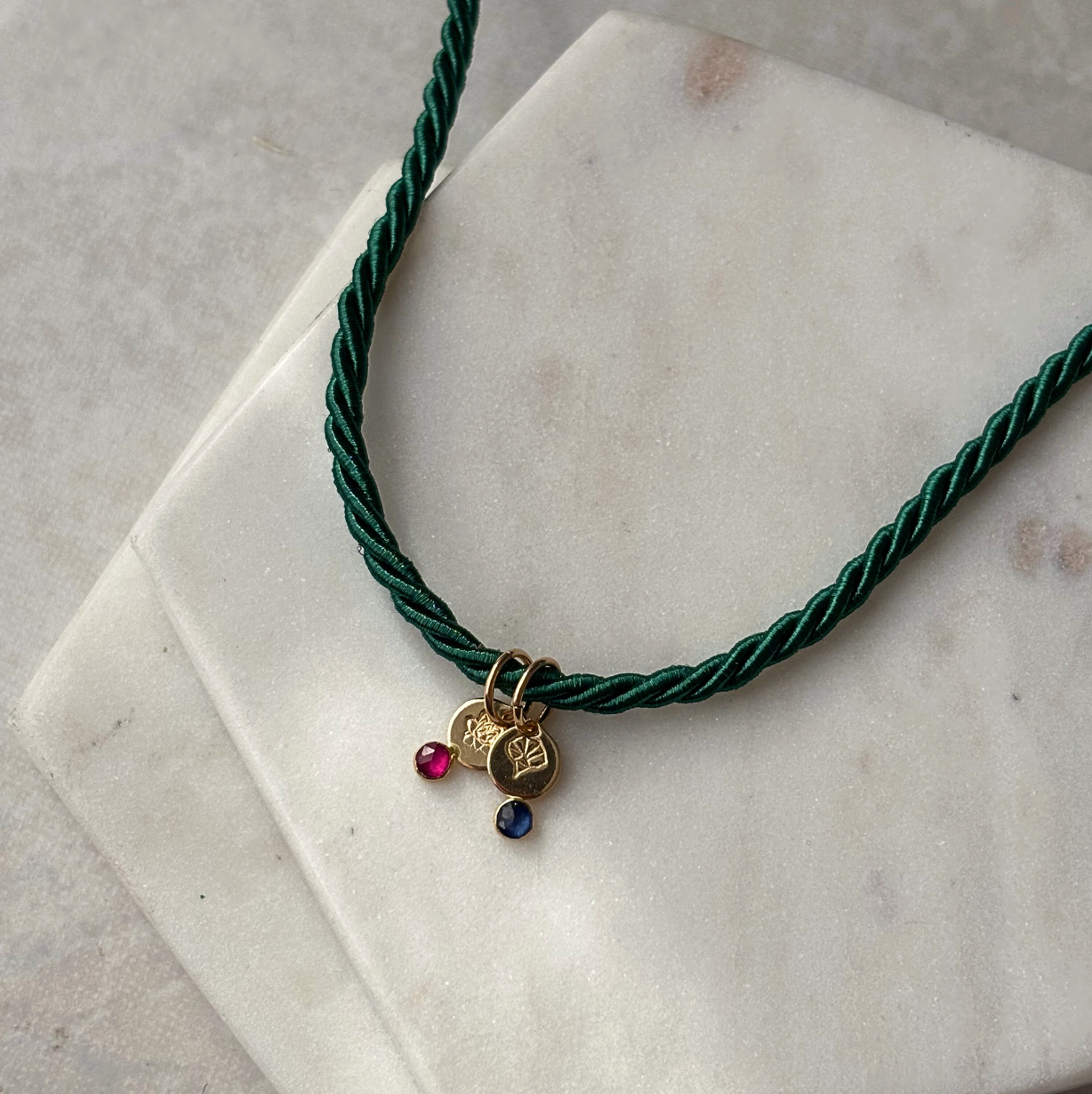 Green cord necklace with gold charm and colored stones on a marble surface
