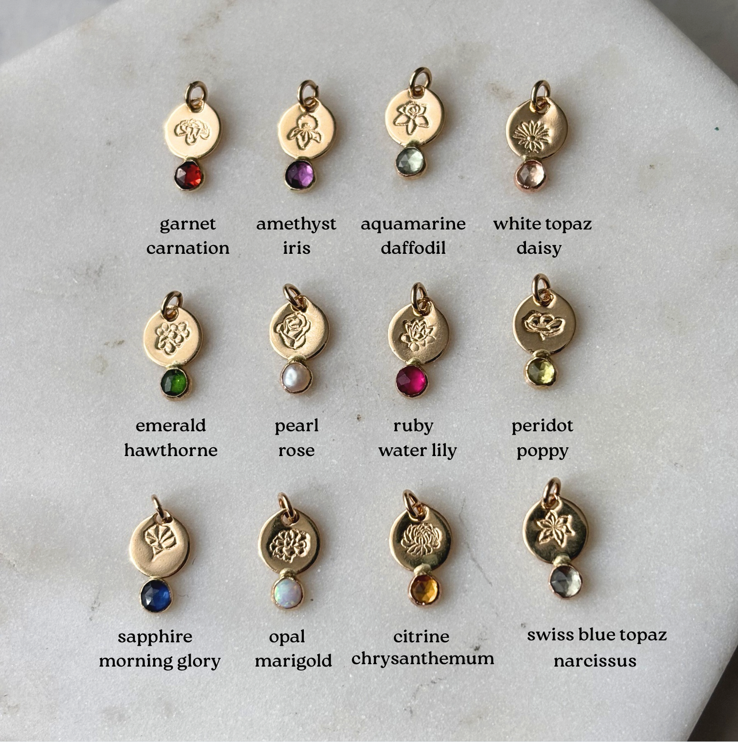 Collection of gold charms with gemstones and corresponding birthstone names on a marble surface