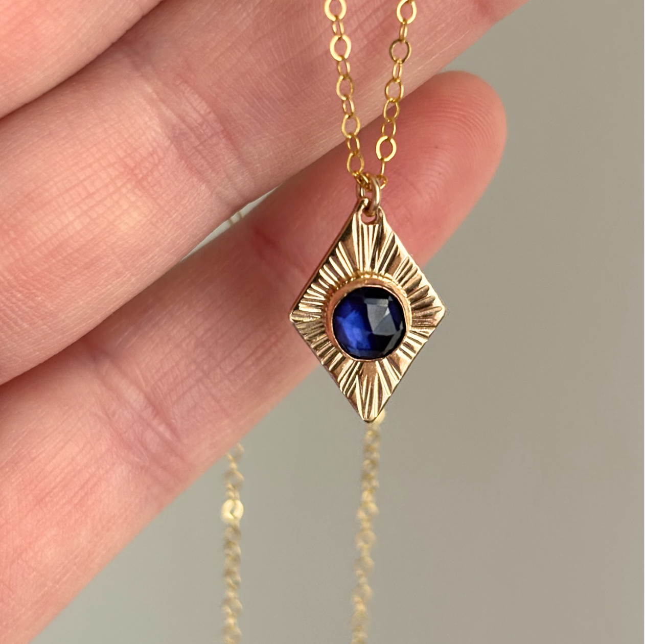 Gold necklace with a blue gemstone pendant held by a hand against a neutral background