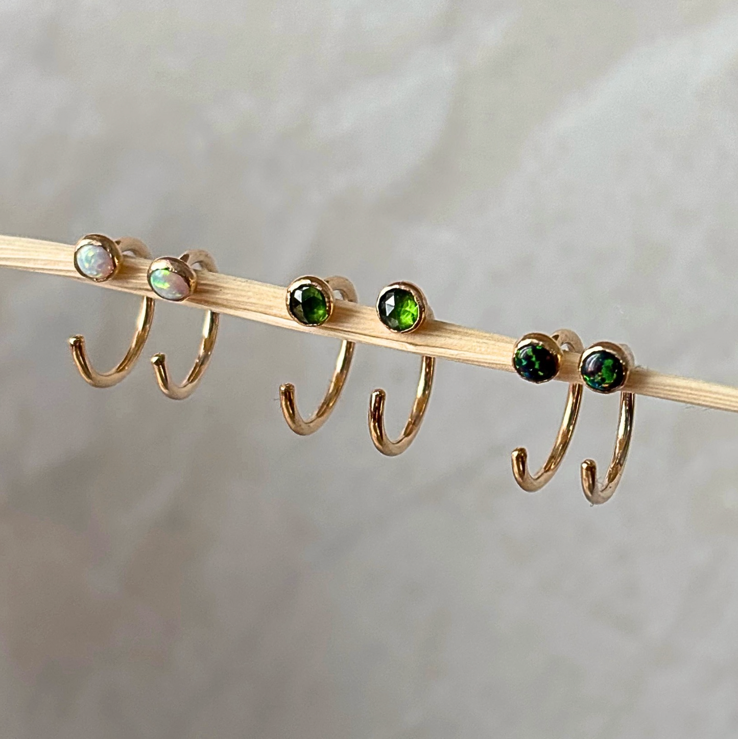 Four gold hoop earrings with round gemstones are displayed on a wooden stick against a soft, light gray fabric background. Two earrings feature iridescent white opal-like stones, and two feature deep green stones.