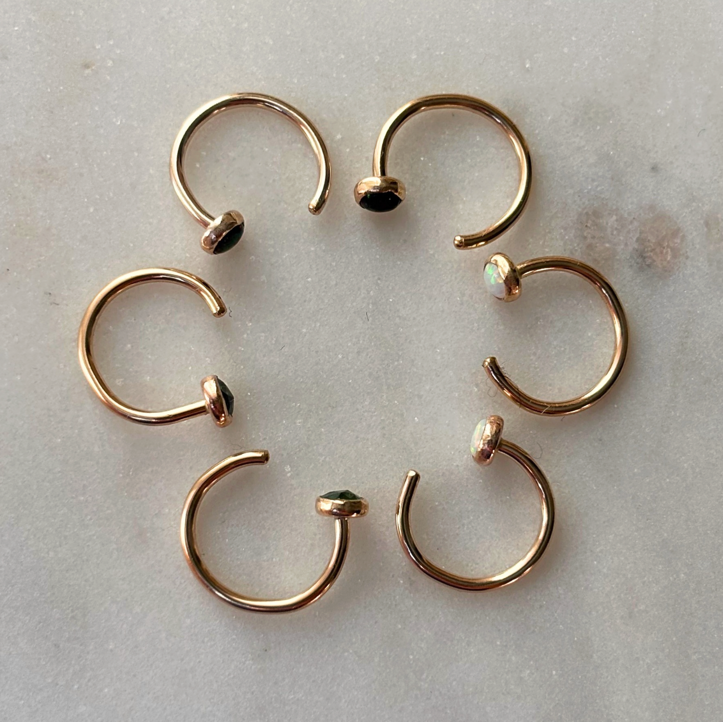 Six gold open hoop earrings with small gemsotne cabochons, arranged in a circle on a light gray marble surface.