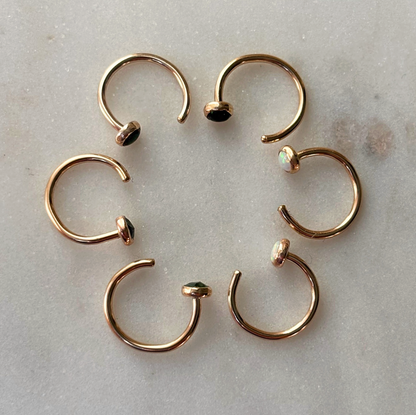 Six gold open hoop earrings with small gemsotne cabochons, arranged in a circle on a light gray marble surface.