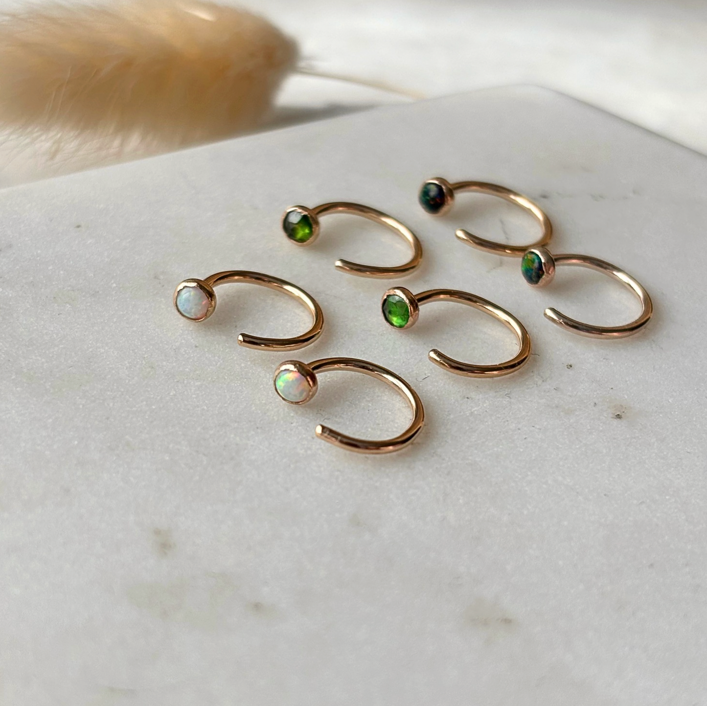 Six gold open hoop earrings with small iridescent gemstone accents in green, blue, and white, arranged on a light marble surface.