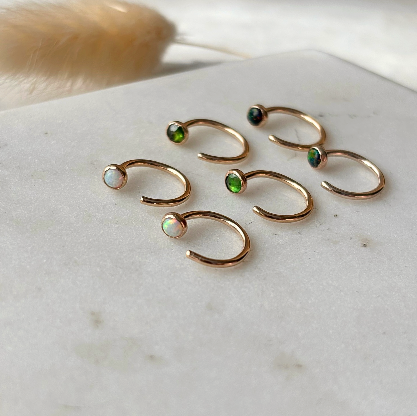 Six gold open hoop earrings with small iridescent gemstone accents in green, blue, and white, arranged on a light marble surface.
