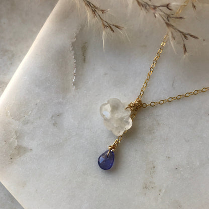 A rainbow moonstone cloud pendant with a purple tanzanite teardrop, displayed on a marble background.