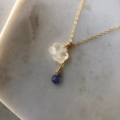 A rainbow moonstone cloud pendant with a purple tanzanite teardrop, displayed on a marble background.