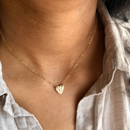 A person wearing a gold heart-shaped textured pendant necklace, displayed against a casual white shirt background.