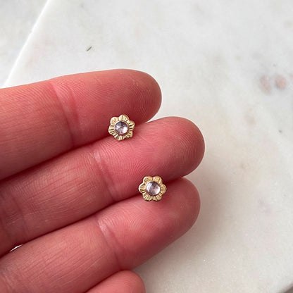 A hand holding a pair of mini flower stud earrings with textured details and tanzanite gemstones.