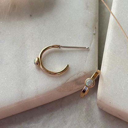 A gold crescent-shaped hoop earring with a small iridescent stone and a silver post, displayed next to a gold ring with an oval white opal on a light stone surface.