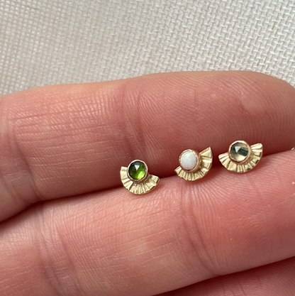 Three small gold earrings with gemstones held between fingers on a light fabric background