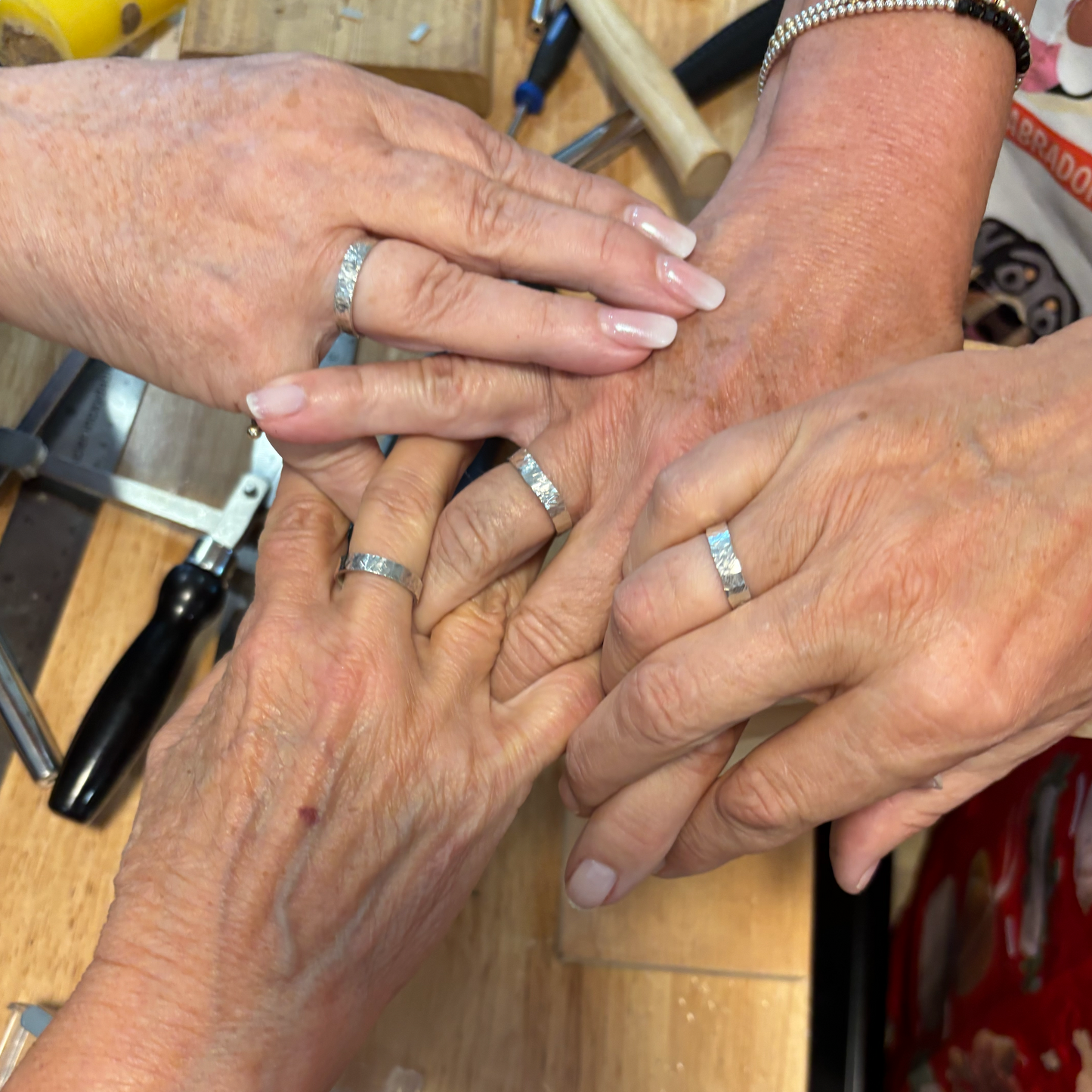 Close-up of four hands with wide silver band rings with jewellery making tools.